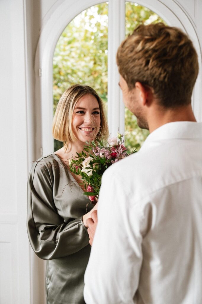 Et yngre par udveksler blomster i en hyggelig indendørs indstilling på deres bryllupsdag. Kvinden, iført en elegant, grøn kjole, smiler bredt, mens hun modtager en smuk buket fra manden, der er klædt i en hvid skjorte. De står foran et stort vindue, som bringer naturligt lys ind i rummet.