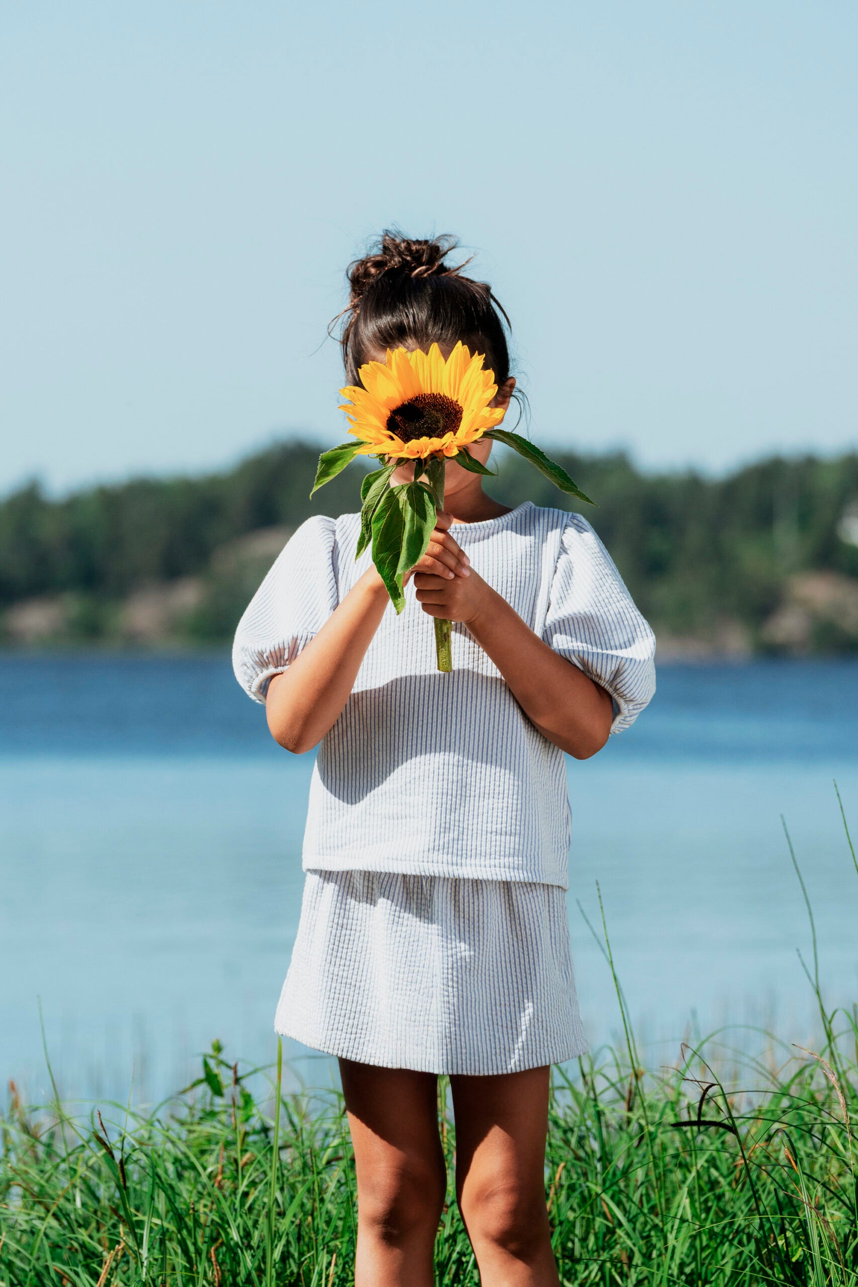Ung pige med en stor gul solsikke i hænderne, smilende i naturligt lys – blomster til Mors Dag fra Interflora