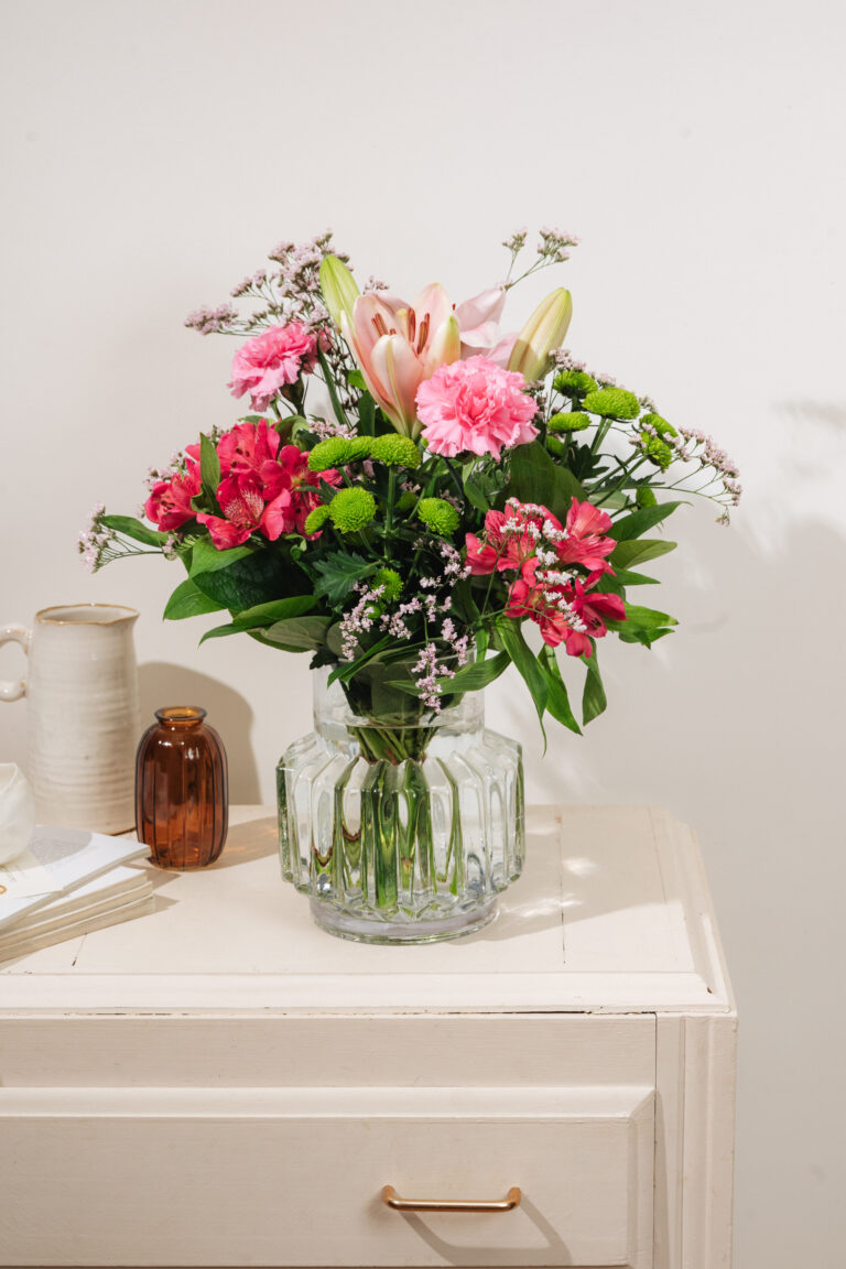 Interflora-buketten Den fine liljehilsen i pink nuancer, placeret i vase på en hvid kommode med skandinavisk indretning