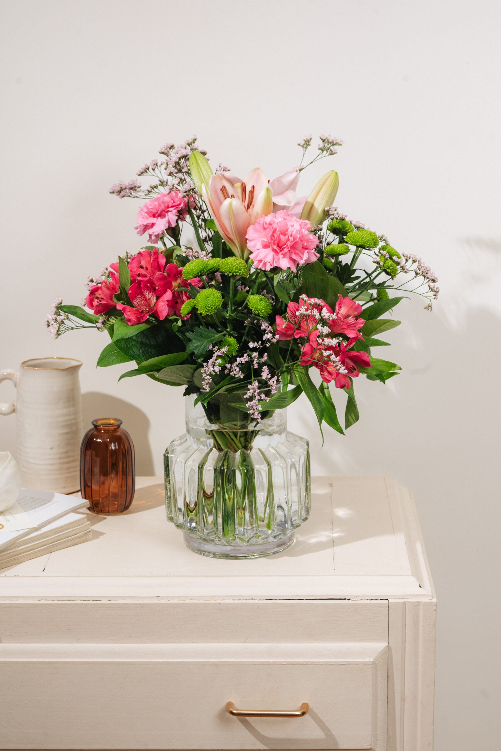 Interflora-buketten Den fine liljehilsen i pink nuancer, placeret i vase på en hvid kommode med skandinavisk indretning