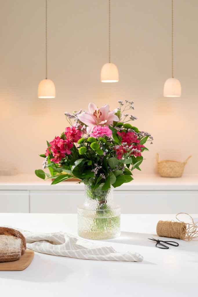Interflora-buketten Den fine liljehilsen med lyserøde liljer og grønne krysantemum i vase på et skandinavisk køkkenbord