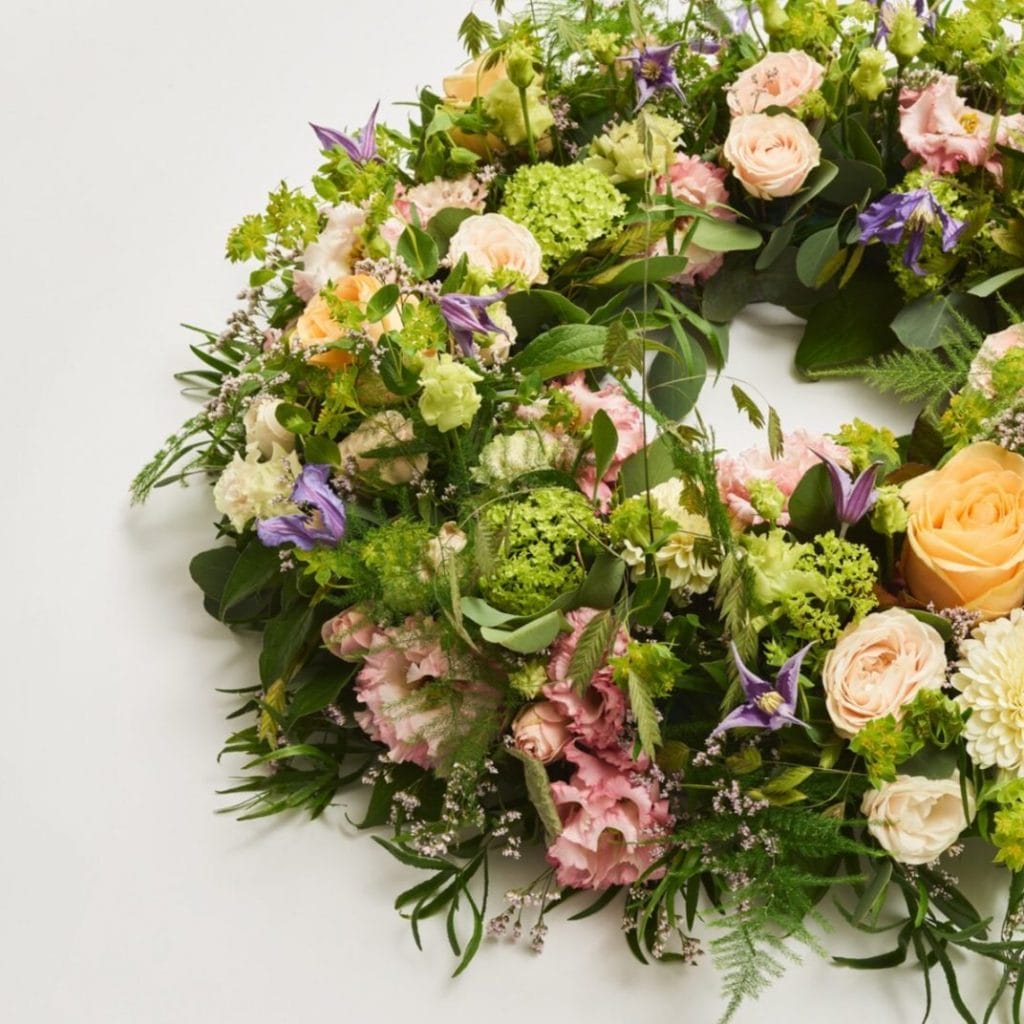 En anden blomsterkrans med en blanding af pastelrosa, lilla, gule og hvide blomster. Kransen er omkranset af grønt og forskellige blomster, som giver et blidt og fredfyldt udtryk. Denne krans bruges til at ære og mindes den afdøde ved en begravelse.