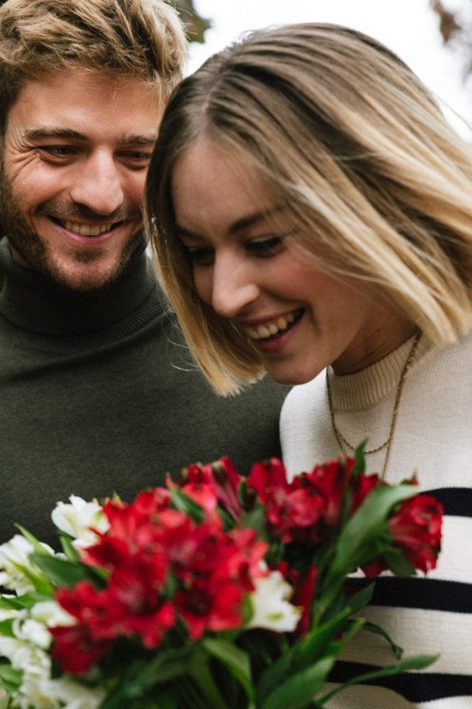 Samme yngre par ses her udendørs på deres bryllupsdag, hvor de begge smilende ser på en buket af røde og hvide blomster. Manden, i en grøn sweater, står tæt ved kvinden, der har en stribet trøje på. De ser ud til at nyde et intimt øjeblik sammen i en frodig have eller park.