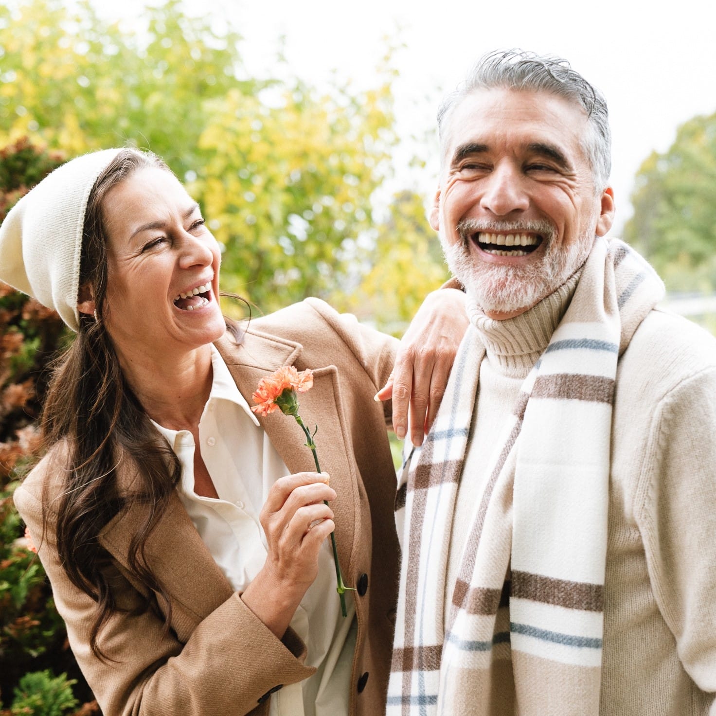 Et ældre par fejrer deres bryllupsdag med store smil. Kvinden holder en orange nellike i hånden, og manden har en stribet halstørklæde og en beige sweater på. De står tæt sammen og deler et øjeblik af glæde og latter i en smuk, grøn park på deres bryllupsdag.