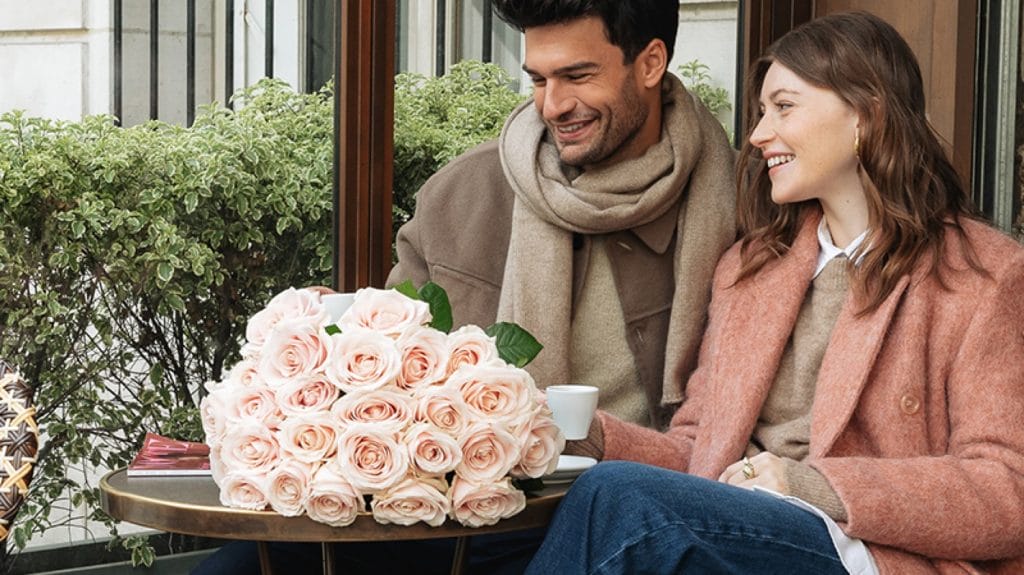 Et par sidder på en caféterrasse. På bordet står en buket lyserøde roser, som er en gave til Valentinsdag.