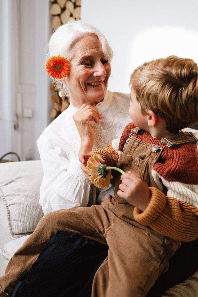 bedstemor med blomst i håret griner sammen med barnebarn på skødet med blomst i hånden